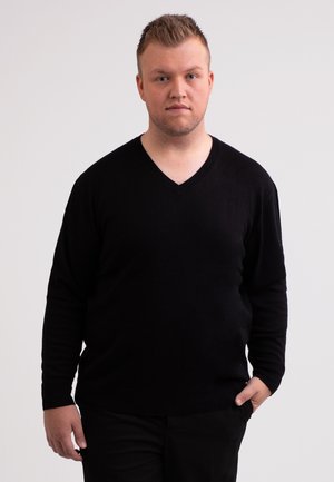 Suéter negro de cuello en forma de V hecho de material suave, con mangas largas y un diseño ajustado. El modelo se encuentra de pie contra un fondo neutro.