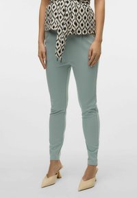 Helle tealfarbene, dehnbare Hosen mit slim fit, kombiniert mit einem gemusterten, mit Gürtel versehenen Oberteil, das schwarze und cremefarbene Rautenformen zeigt. Schuhe in neutralen Farben.