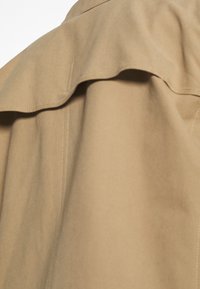 Béžová trenčkotová tkanina s detailním skláp ěcím límcem. Hladký povrch a jemný lesk, který zdůrazňuje švy a štepování.