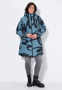 Giacca esterna impermeabile color teal con motivo astratto nero, chiusura con zip, cappuccio regolabile e maniche lunghe, abbinata a pantaloni grigi e stivali neri.