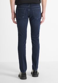 Hombre que lleva unos pantalones vaqueros oscuros ajustados y zapatos de cuero negros, de pie contra un fondo blanco liso, visto desde detrás.