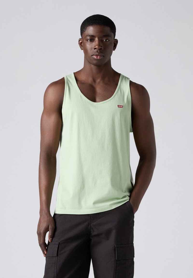 Jonge man staat met één hand in zijn zak, draagt een lichtgroen hemdje en zwarte cargobroek tegen een effen lichte achtergrond.