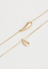 Divi zelta kaklarotas ar gludām, neregulāras formas piekariņiem uz tievām, pulētām ķēdēm uz vienkārša balta fona.