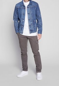 Mies, który ma na sobie niebieską dżinsową kurtkę, białą koszulkę, szare spodnie cargo i białe sneakersy, stoi na jednolitym szarym tle.