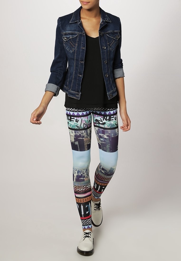 Femme portant une veste en jean foncé, un haut noir, des leggings colorés à motifs de paysages urbains, et des chaussures blanches à lacets, debout sur un fond clair.