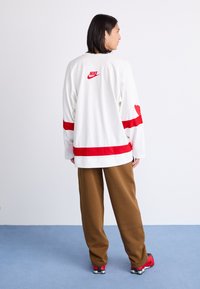 En person med langt mørkt hår står sideværts iført en hvid Nike sportsjersey med røde striber, brune bukser og røde sneakers.