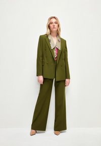Olivgrön skräddarsydd kavaj med dubbelknäppta knappar, breda slag, matchande vida byxor och en beige broderad blus under.