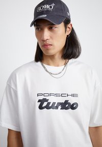 Balta T-kreklu ar lielu melnu "PORSCHE turbo" uzrakstu centrā. Apkakli rotā divas sudraba ķēdes. Tumšs beisbola cepure ar uzrakstu "SOHO MANHATTAN."