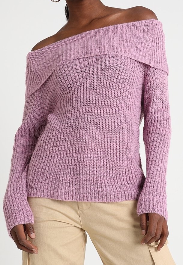 Femme portant un pull tricoté mauve clair à épaules dénudées avec des manches longues et un pantalon beige, debout devant un fond uni.
