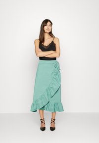 Saia envolvente verde menta com bainha em babados, feita de tecido suave. Acompanhada de uma camisola preta com rendas e saltos pretos com tiras.