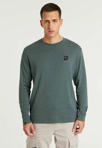 Langärmliges grünes Hemd aus weichem Material, mit einem kleinen schwarzen Logo-Patch auf der Brust und gerippten Bündchen.