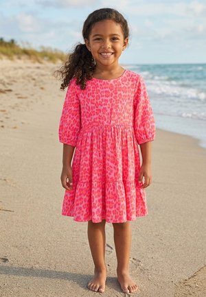 Junges Mädchen lächelt, trägt ein rosa gemustertes Kleid, steht barfuß an einem Sandstrand mit Meereswellen im Hintergrund.