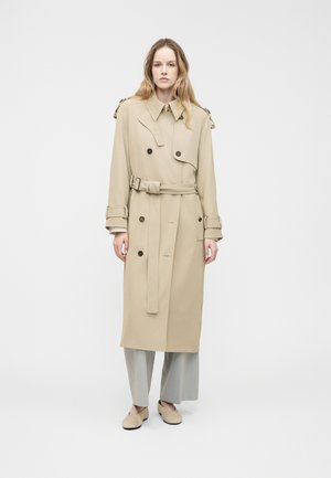 Kvinde med langt hår iført en beige trenchcoat med bælte, lysegrå bukser med vide ben og beige slip-on sko, stående mod en ensfarvet baggrund.
