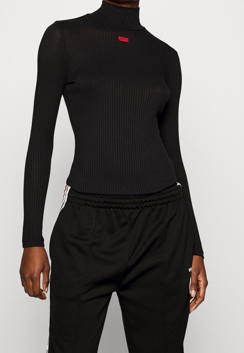 Personne portant un col roulé côtelé noir avec un petit logo rouge sur la poitrine et un pantalon noir à taille élastique avec des bandes blanches et rouges.