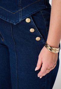 Donkerblauwe denim broek heeft gouden knoopaccenten aan de zijkant en contrasterende stiksels, weergegeven naast een hand versierd met ringen en armbanden.
