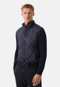 Gilet matelassé bleu marine avec un col haut et une fermeture éclair, associé à des manches longues bleu marine. Il comprend deux poches zippées sur les côtés et un ourlet côtelé.
