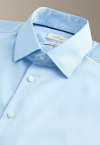 Šviesiai mėlyna ilgomis rankovėmis palaidinė su sagomis ant apykaklės, penkiomis baltomis sagutėmis ir kontrastingų spalvų vidiniu kaklo ženklinimu.