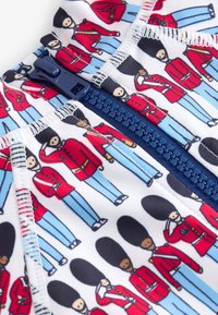 Badebekleidungsstoff mit einem sich wiederholenden Muster von Cartoon-Wächtern in roten Uniformen und blauen Hosen, mit einem marineblauen Reißverschluss und weißem Hintergrund.