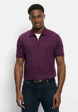 Mann trägt ein dunkelviolettes kurzärmeliges Poloshirt mit einem vorderen Reißverschluss, dunkle Jeans und einen schwarzen Gürtel, stehend vor einem einfarbigen Hintergrund.