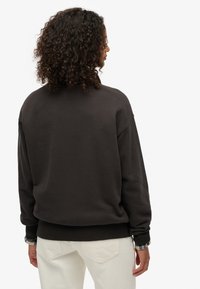 Felpa marrone scuro con vestibilità rilassata, collo rotondo e polsini a costine. Texture morbida; indossata sopra una camicia a quadri chiari, con pantaloni color crema.