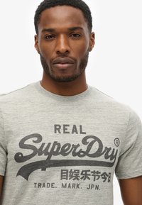 Camiseta gris de manga corta hecha de tejido texturizado que presenta un gran logo impreso en negro que dice "REAL SuperDry" y texto adicional debajo.