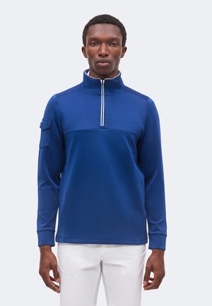Homme portant un haut zippé bleu à manches longues avec un col haut et une poche sur la manche gauche, associé à un pantalon blanc sur un fond uni.