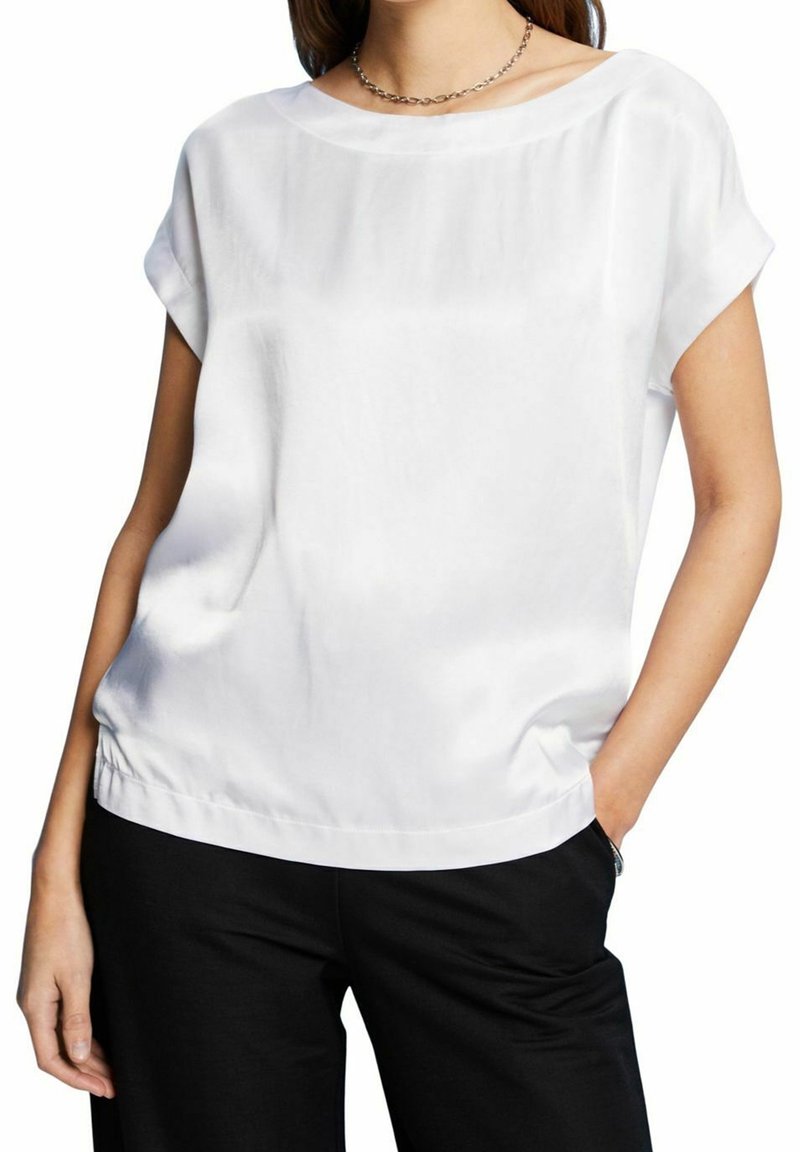 Femme portant un chemisier blanc ample à manches courtes et un pantalon noir, une main dans la poche et un collier chaîne en argent.