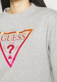 Šedá mikina s velkým trojúhelníkovým logem s gradientními barvami oranžové a růžové, na kterém je nápis "GUESS" a "WASHED JEANS".