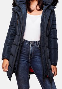 Mörkblå vadderad jacka med pälsfodrad huva, som har dragkedja framtill, sidfickor och ett quiltat mönster. Bärs över en vit topp och jeans.