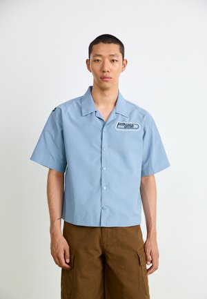 Short-sleeve light blue shirt with a button-up front, featuring a black and white Puma logo on the chest and a relaxed fit design.