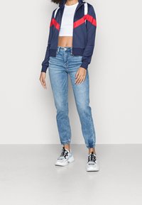 Žena v námořnickém sakě s červenými pruhy, bílém crop topu, světle modrých teplákových kalhotách a bílých teniskách stojící na jednobodovém pozadí.