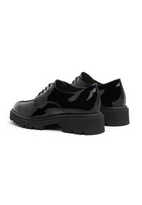 Zapatos de charol negro con punta redondeada, suela gruesa tipo lug y costura decorativa a lo largo del borde de la suela. Presenta ocho ojales para los cordones.