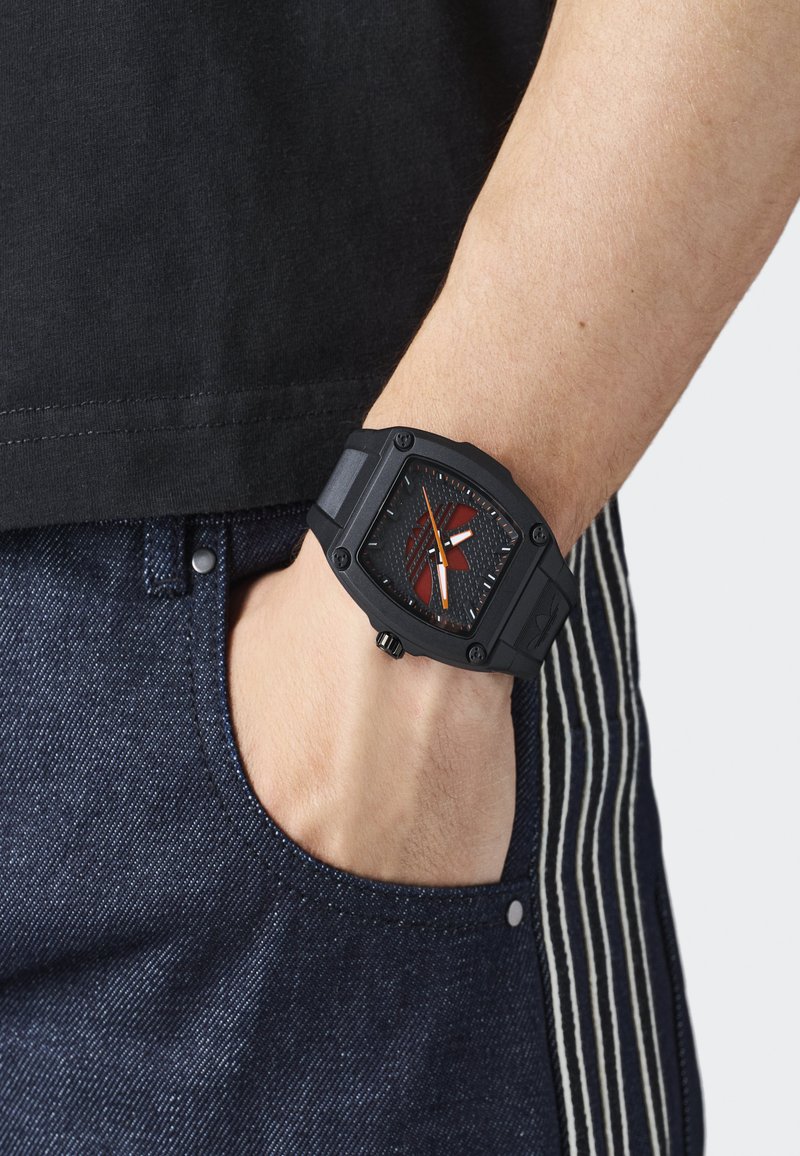 Osoba nosząca czarny zegarek na nadgarstku o kwadratowej tarczy z pomarańczowymi akcentami, ręka swobodnie umieszczona w kieszeni ciemnoniebieskich dżinsów.