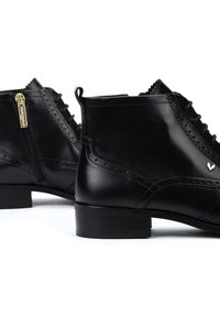 Botas de cuero negro hasta el tobillo con punta redonda, detalles de broguing decorativos, cremallera dorada y un tacón bajo y cuadrado. Textura suave con acentos de costura.