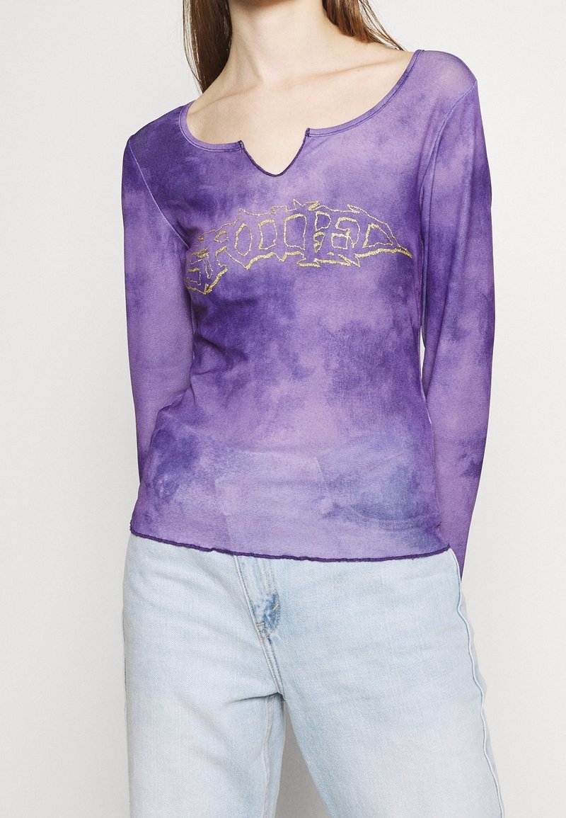 Fialový top s dlouhými rukávy a kulatým výstřihem, zdobený zlatým grafickým designem. Materiál má efekt batiky a mírně průsvitnou texturu.