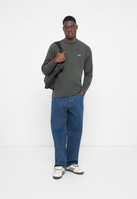 Langermet mørkegrønn skjorte med logo, kombinert med overdimensjonerte blå denimjeans og hvite joggesko med svarte striper. Svart veske slengt over skulderen.