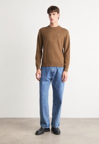 Bruine gebreide trui met lange mouwen, ronde halslijn en een klein logo; gecombineerd met lichtblauwe rechttoe-rechtaan jeans en zwarte schoenen.