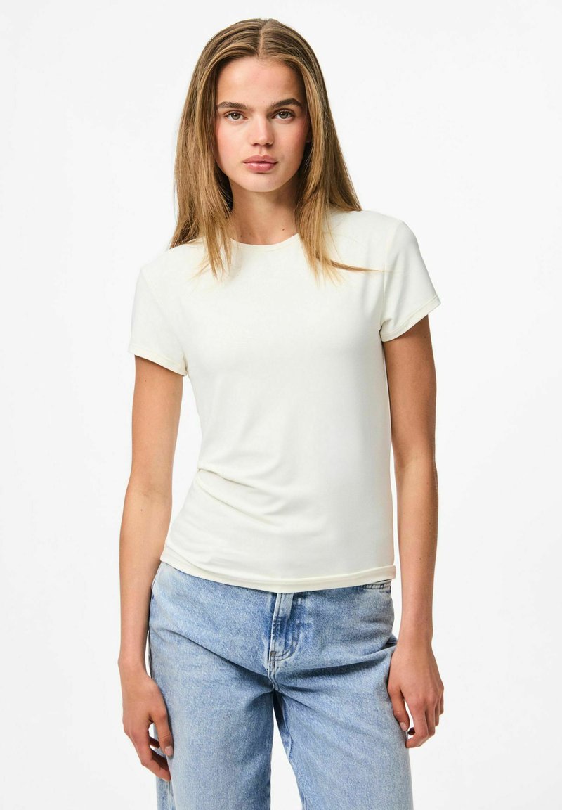 Kreker krāsas kokvilnas auduma T-krekls ar īsām piedurknēm un apaļu kakla izgriezumu, pieguļošs dizains, gluda tekstūra, kopā ar gaiši ziliem džinsiem.