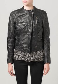 Schwarze Lederjacke mit einem cropped Design, ausgestattet mit Reißverschluss-Taschen, einem Stehkragen und einer strukturierten Oberfläche. Getragen über einer gepunkteten Bluse.
