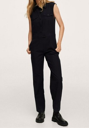 Kvinde iført en ærmeløs sort jumpsuit med knappede brystlommer og sorte robuste ankelstøvler, stående med hænderne i lommerne.