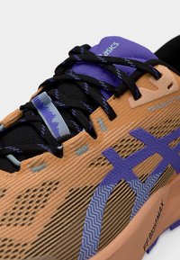 Τα αθλητικά παπούτσια Asics Trabuco 14 διαθέτουν πορτοκαλί και μαύρο υφασμάτινο άνω μέρος, μοβ λεπτομέρειες και μαύρα κορδόνια με ζιγκ-ζαγκ μοτίβο.