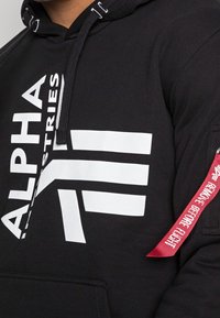 Felpa con cappuccio nera caratterizzata da un grande logo bianco "ALPHA" e tre strisce orizzontali bianche. Etichetta di tessuto rossa sulla manica con la scritta "RIMUOVERE PRIMA DEL VOLO."
