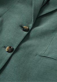 Gros plan d'une veste en velours côtelé verte avec deux boutons en écaille et une poche fendue sur le panneau avant.