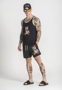 Čierny tielko s viacfarebnou grafikou medveďa, doladené s čiernymi šortkami s podobným dizajnom medveďa, doplnené o čierne šľapky.