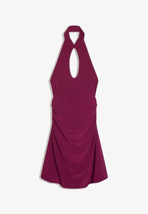 Junge Frau, die ein ärmelloses violettes Neckholderkleid mit Schlüssellochausschnitt und bunte Armreifen trägt, steht nahe bei Strandfelsen und Ozean.