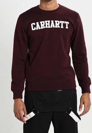 Mand iført en mørkerød sweatshirt med hvid "CARHARTT"-tekst, sorte bukser og en sort forklæde-stil tilbehør med hvide stropper.
