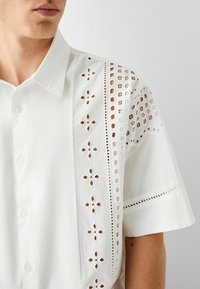 Bílá košile s krátkým rukávem, která má složité vyřezávané vzory podél levého ramene, s knoflíkovým zapínáním vpředu a texturovaným finišem látky.