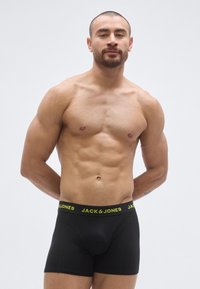 Boxers noirs avec une ceinture élastique jaune affichant la marque « JACK & JONES » ; le mannequin, de carrure musclée, est debout les bras derrière le dos.
