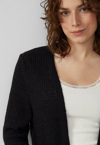 Schwarze strickwarme Strickjacke mit strukturiertem Muster, offenem Vorderteil und langen Ärmeln, über einem weißen gerippten Tanktop mit Spitzenbesatz getragen.
