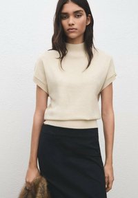 Krämfärgad stickad topp med korta ärmar och hög halsringning, kombinerad med en figurnära svart kjol. Toppen har en lätt struktur och en avslappnad passform.